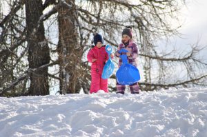 Zwei Kinder im Schnee mit Schaufeln, umgeben von schneebedeckten Bäumen. Winterspaß in den Alpen.